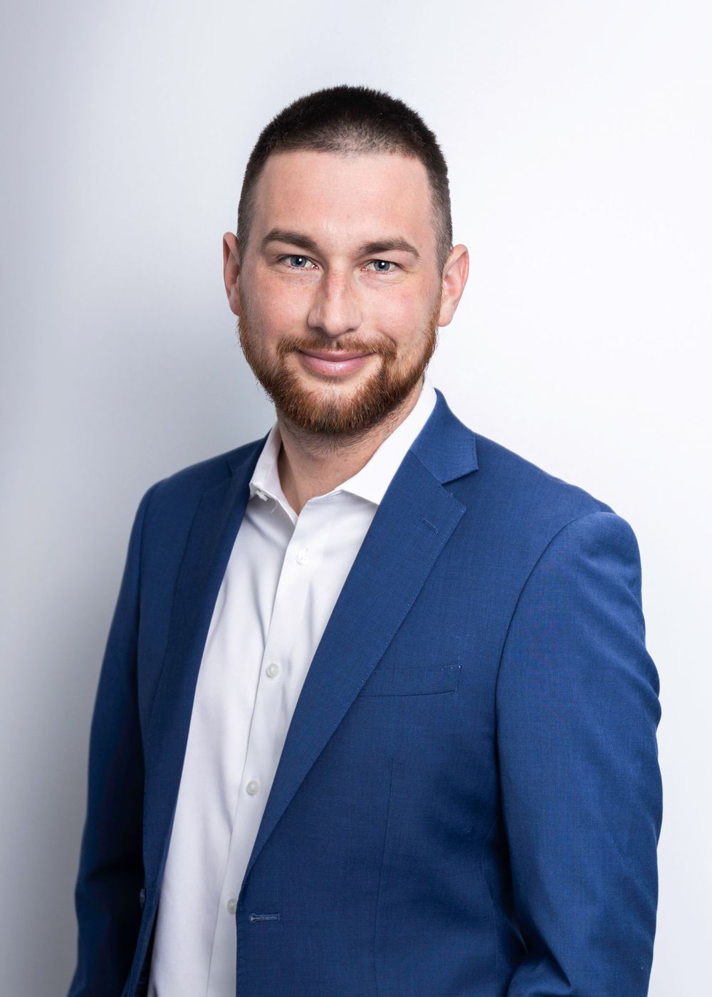 Professional headshot of a person in a navy blue suit jacket and white dress shirt with a well-groomed beard smiling at camera.