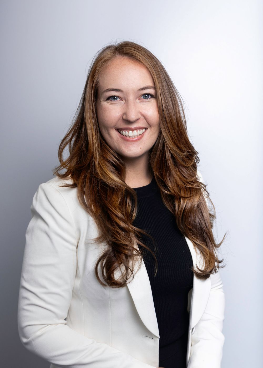 Professional headshot portrait of someone wearing a white blazer and black top against a light gray background, smiling warmly.