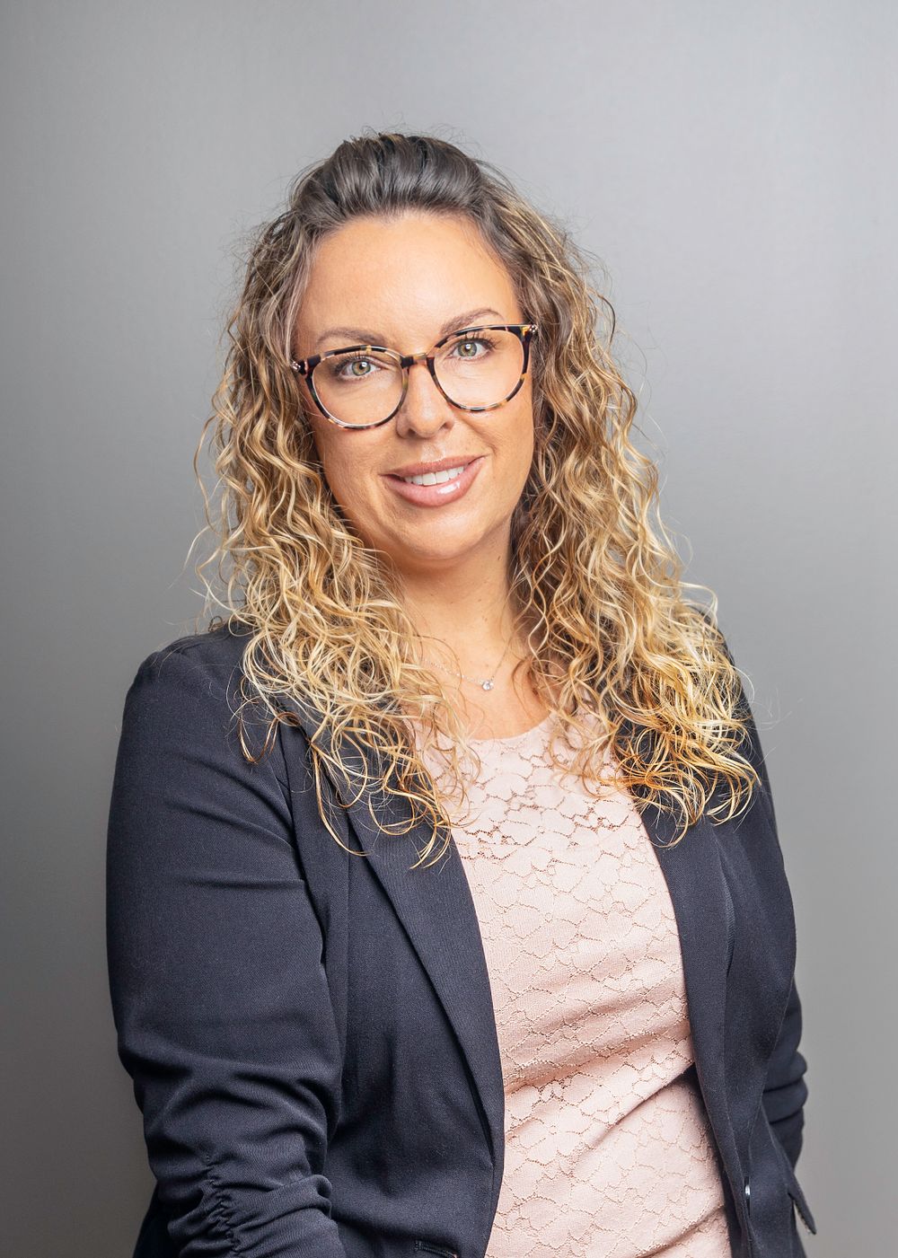 Professional headshot portrait of someone with curly blonde hair wearing glasses, a navy blazer and pink blouse against gray background.