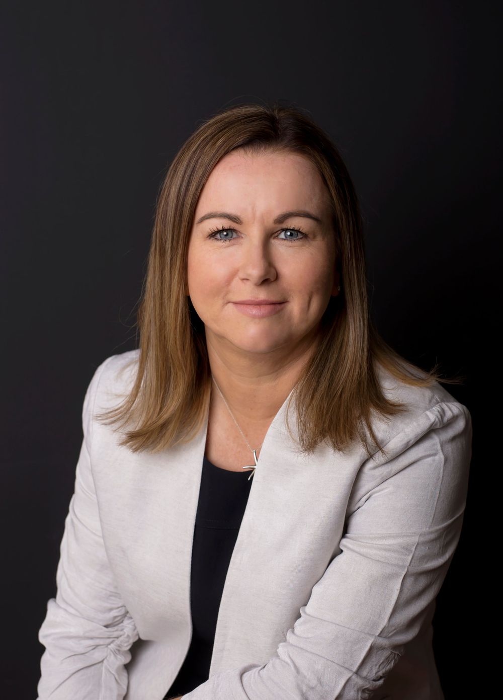 Professional headshot portrait of person in white blazer and dark top against black background.