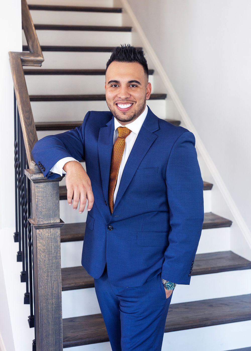 Professional in a well-fitted navy blue suit with orange tie poses confidently on wooden stairs with white walls.