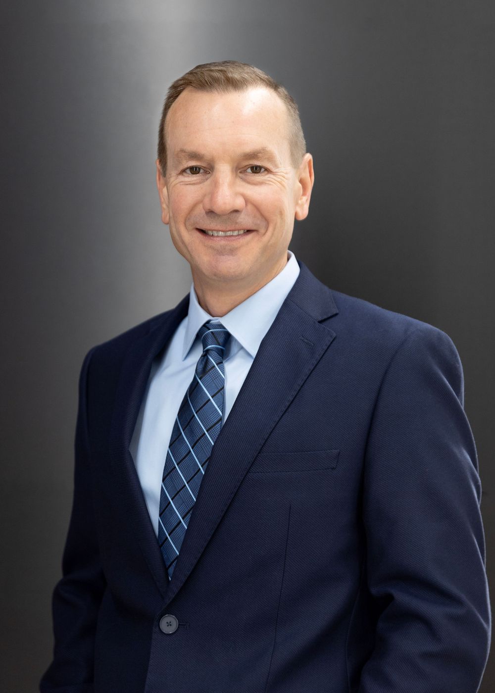 Professional headshot of someone wearing a navy suit and blue plaid tie against a dark background.