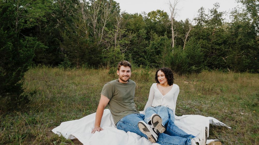 Two people sitting together on a white blanket in a grassy field with trees in the background.