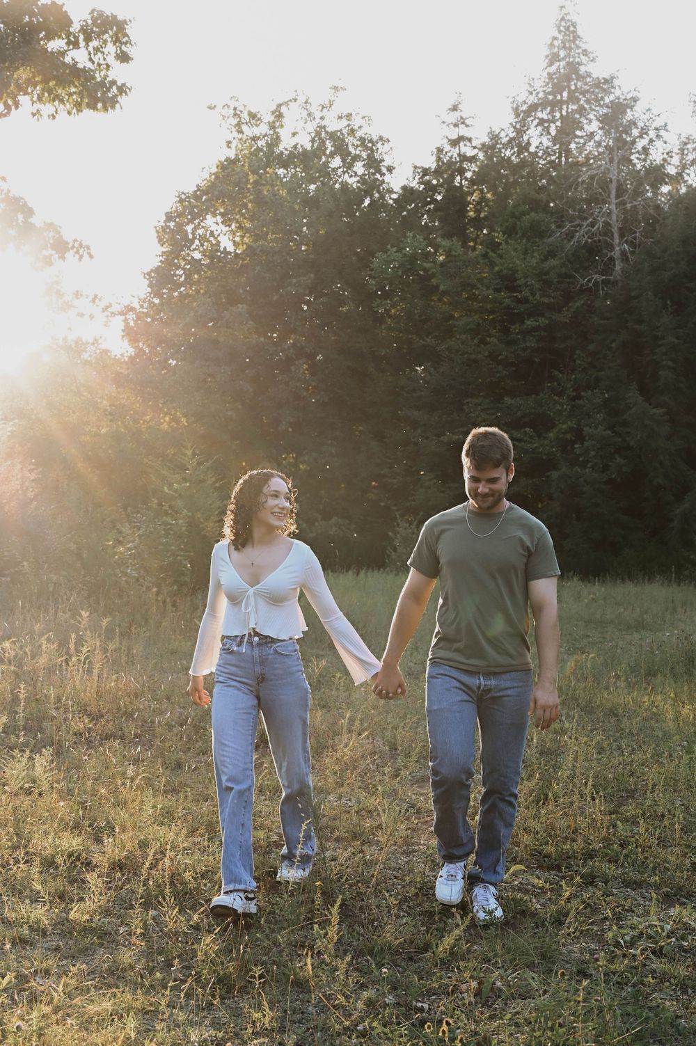A couple walks hand in hand through a grassy field at sunset with sunlight filtering through trees in the background.
