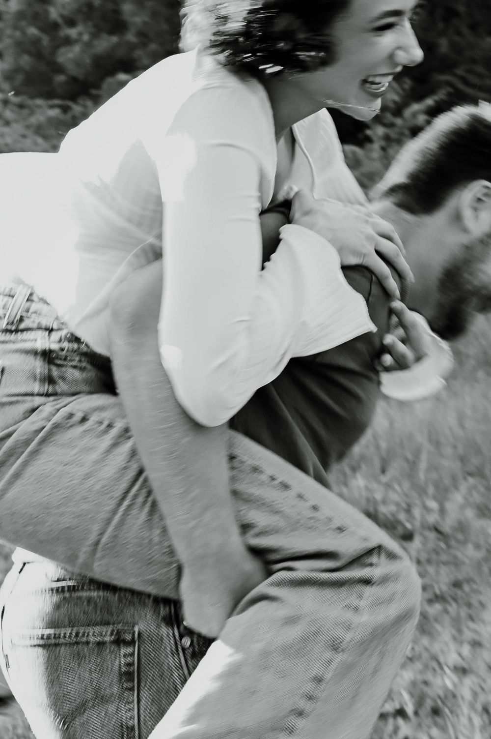 A candid black and white photo showing a couple playfully enjoying a piggyback ride outdoors in vintage casual attire.