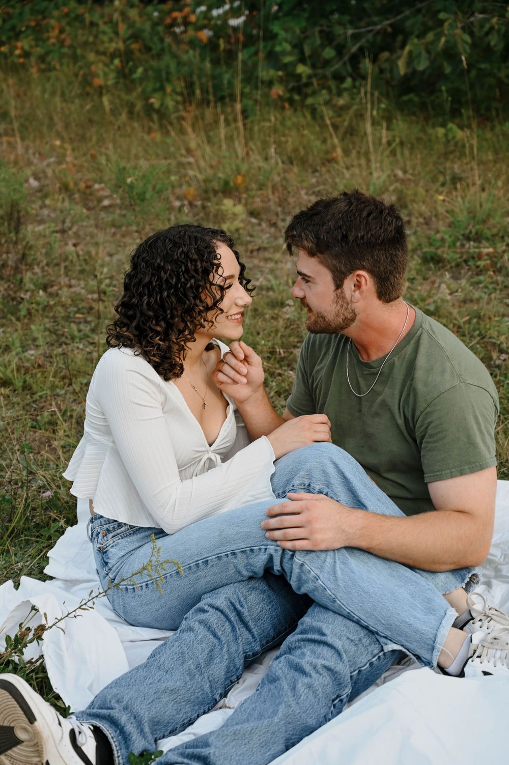 A couple sharing an intimate moment while sitting on a white blanket outdoors in a natural setting.