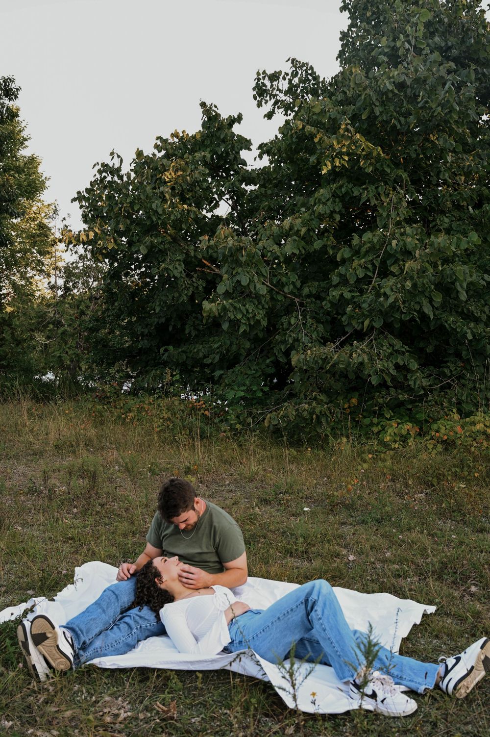 A couple relaxes together on a white blanket in a grassy field with green trees in the background.