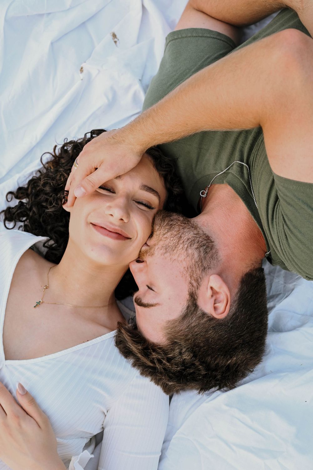 Couple lying down together intimately on white bedding with one wearing an olive green shirt.