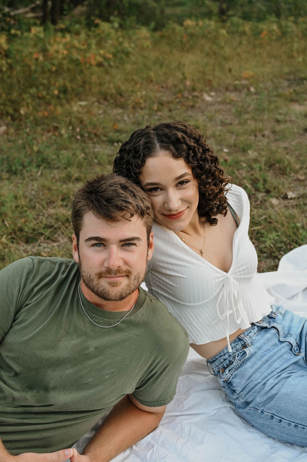 A couple relaxes together on a white blanket outdoors against a natural green background.