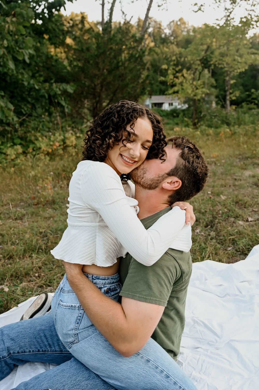 A couple shares an intimate moment while sitting on a white blanket outdoors in a natural setting.