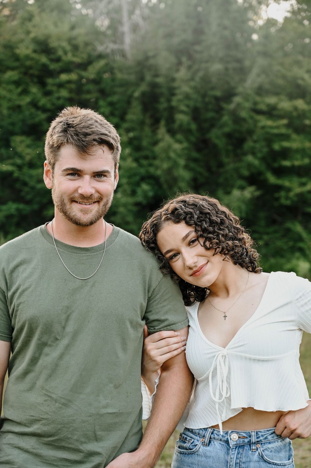 A couple stands close together outdoors against a green forest background on a sunny day.