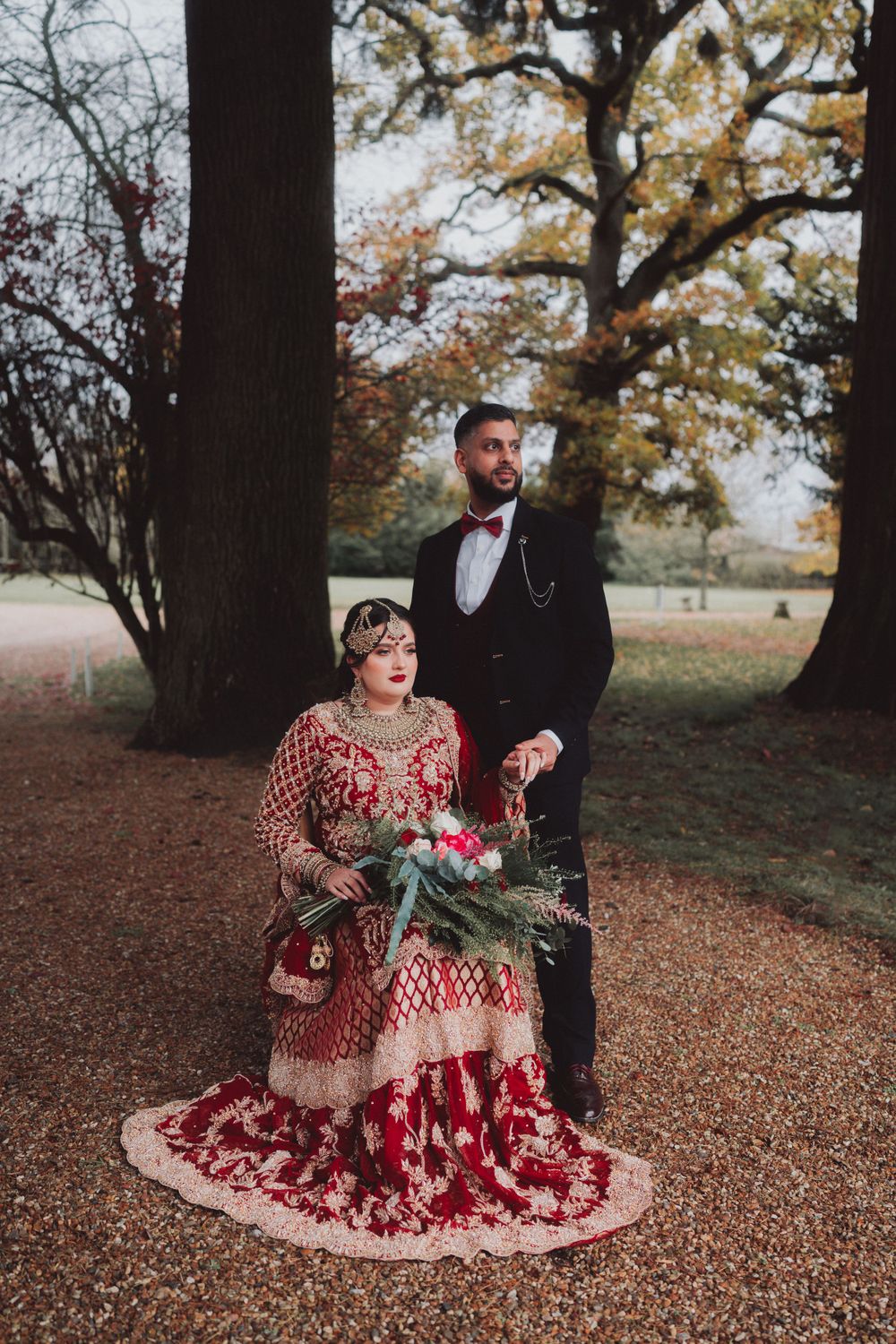 Outdoor wedding portrait featuring a couple in traditional red and black formal attire under autumn trees.