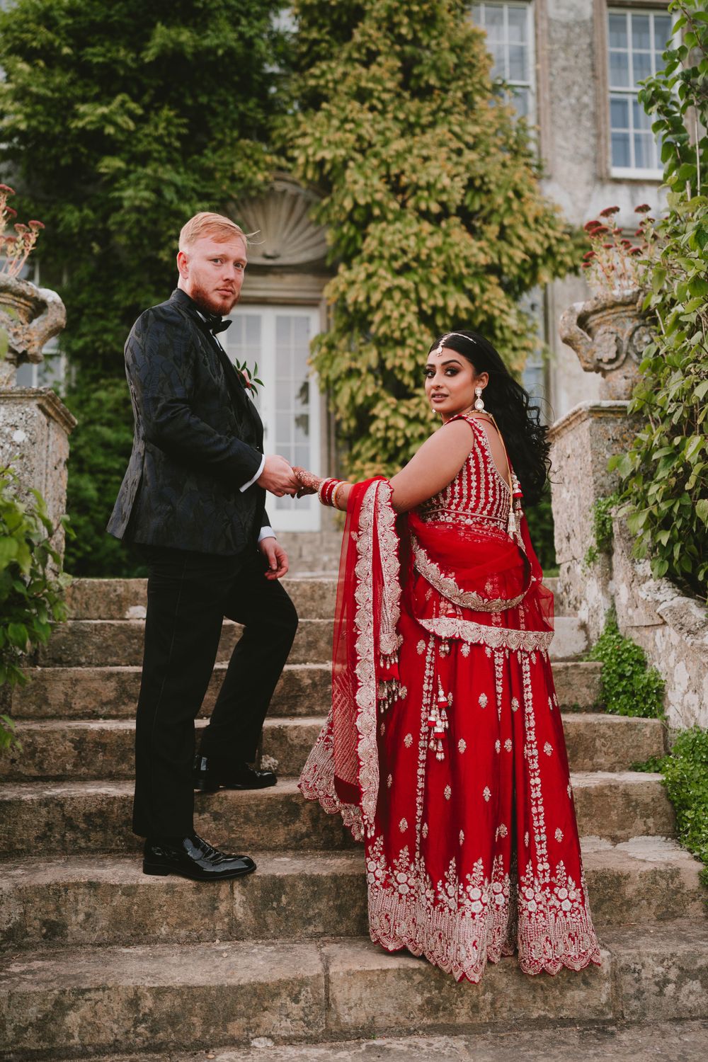 A couple in wedding attire pose on stone steps of an ivy-covered building.
