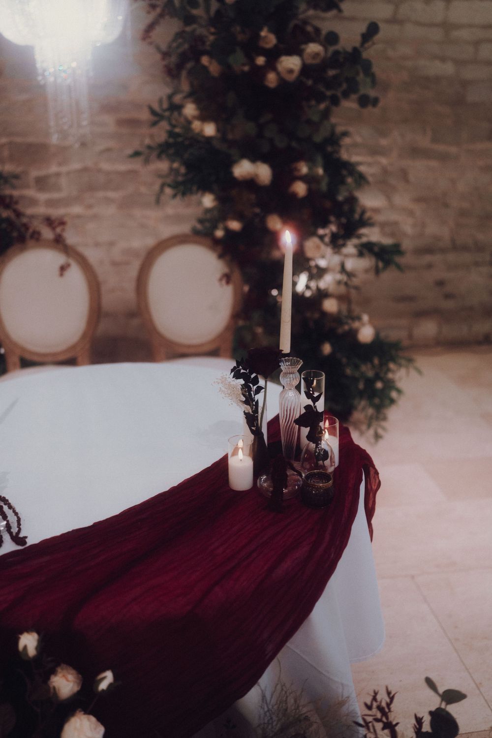 Elegant table setting with burgundy draping and white candles against exposed brick wall.