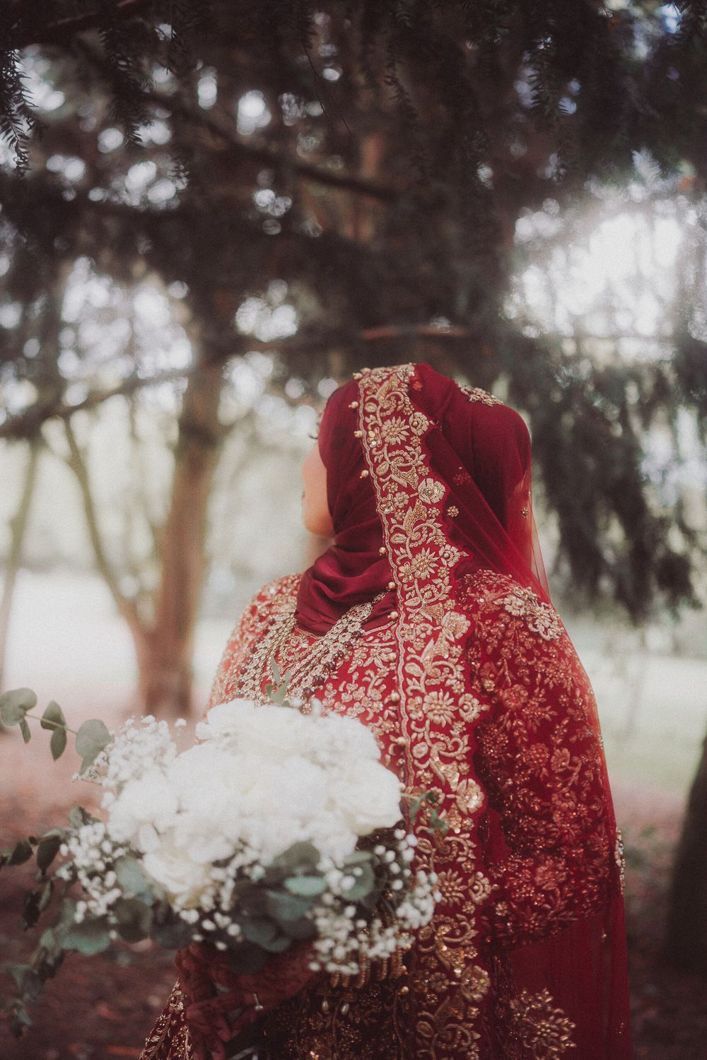 A deep red and gold embellished formal dress or sari with intricate embroidery details in a misty outdoor setting.