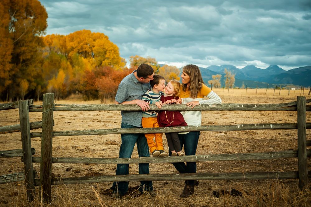 Family wearing autumn colors stands at wooden fence overlooking mountain landscape with golden fall foliage.