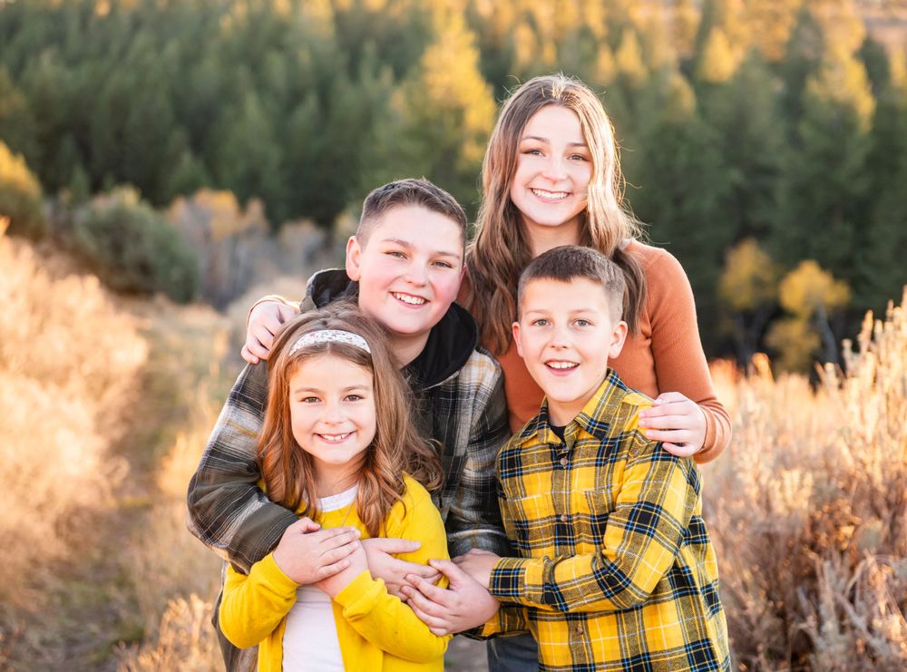 A group of four poses together outdoors against an autumn forest backdrop with golden grass and evergreen trees.
