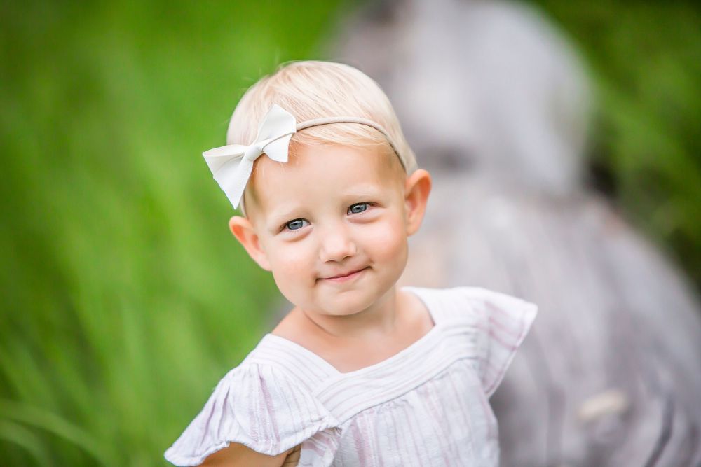 Small child wearing white dress and bow smiles warmly in outdoor garden setting with blurred green background.