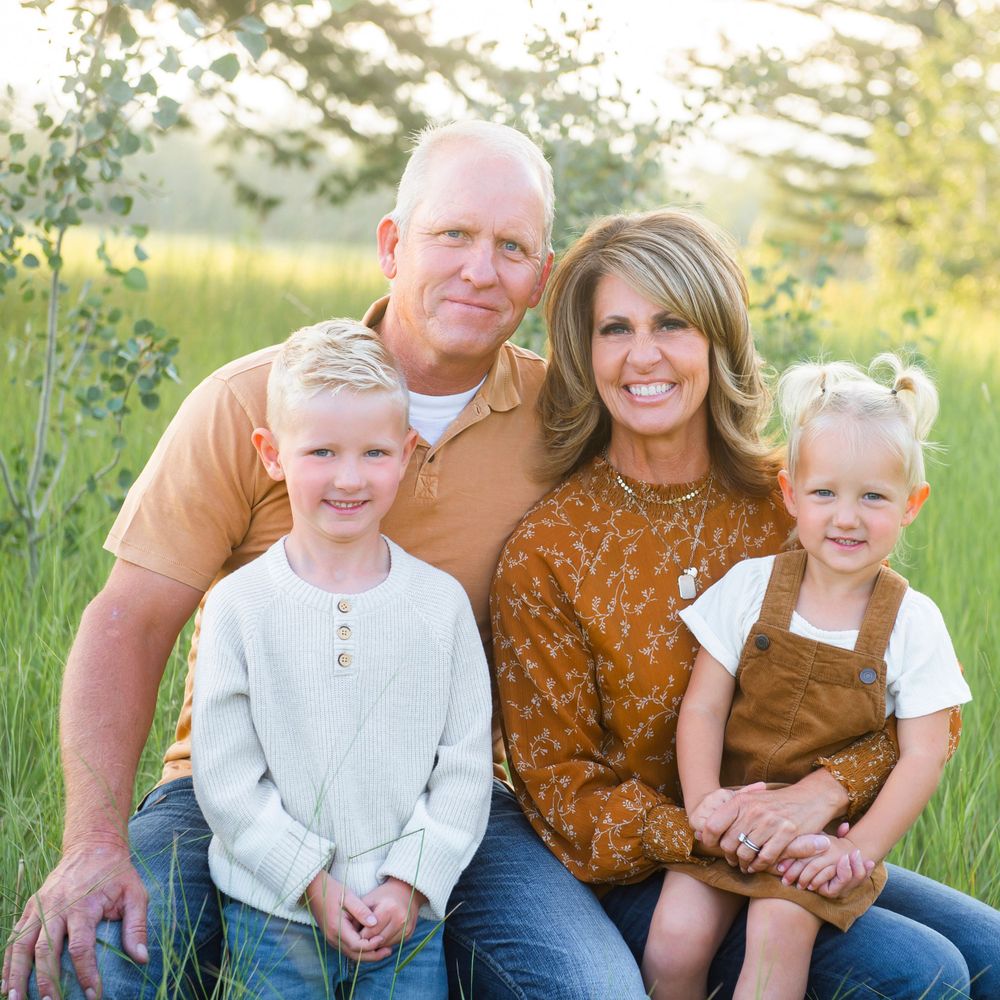 A family of four sits together outdoors in a field during golden hour wearing casual fall attire.