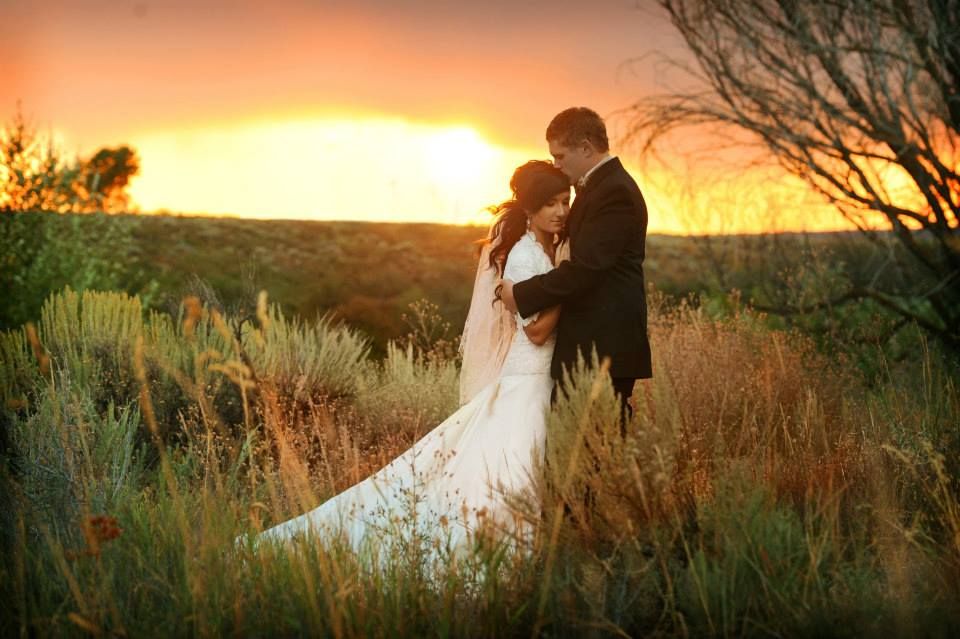 A romantic wedding couple embraces in a desert landscape during a golden sunset with sagebrush and tall grass surrounding them.