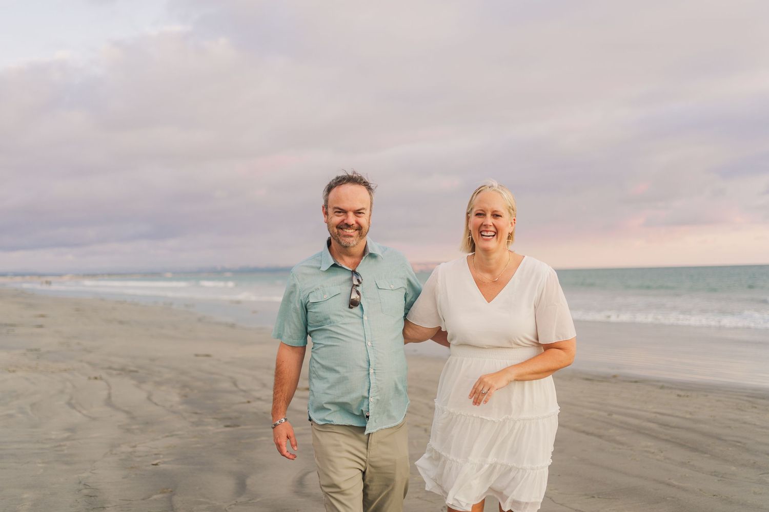 Couple walking on sandy beach at dusk with cloudy sky and ocean waves.