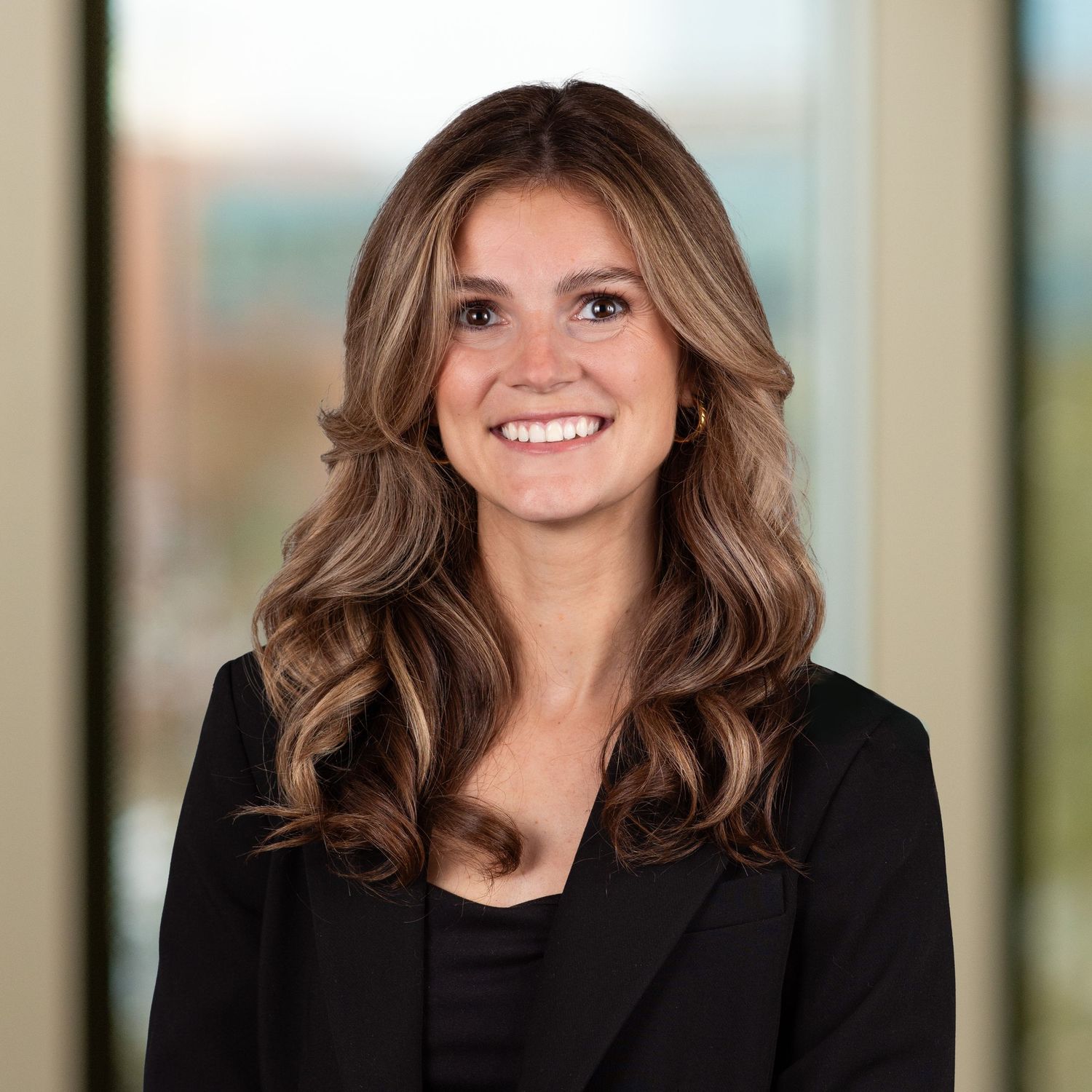 Professional headshot of someone with wavy brown hair wearing a black blazer against a blurred background.