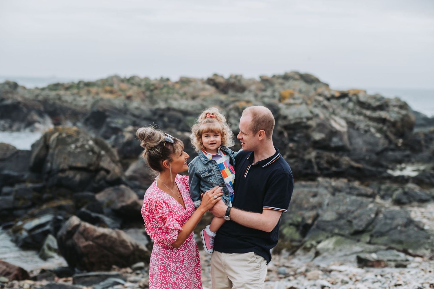 Happy young family with toddler laughing together on a rocky coastal beach shoreline.