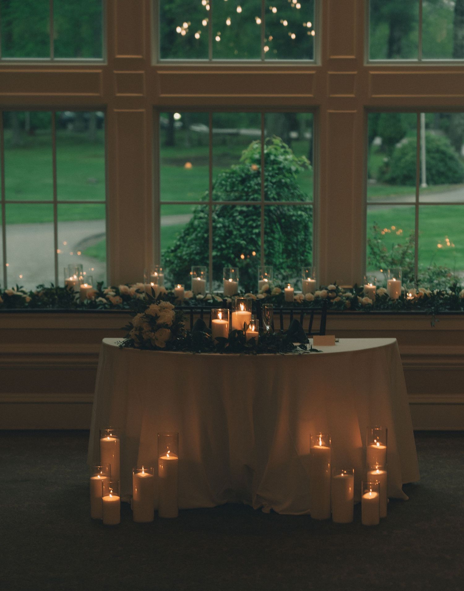 Romantic candlelit table setting with glowing lights and greenery along windowsills at dusk.