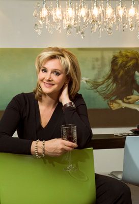 A woman in black attire sits at a table with a glass of wine under an elegant crystal chandelier.