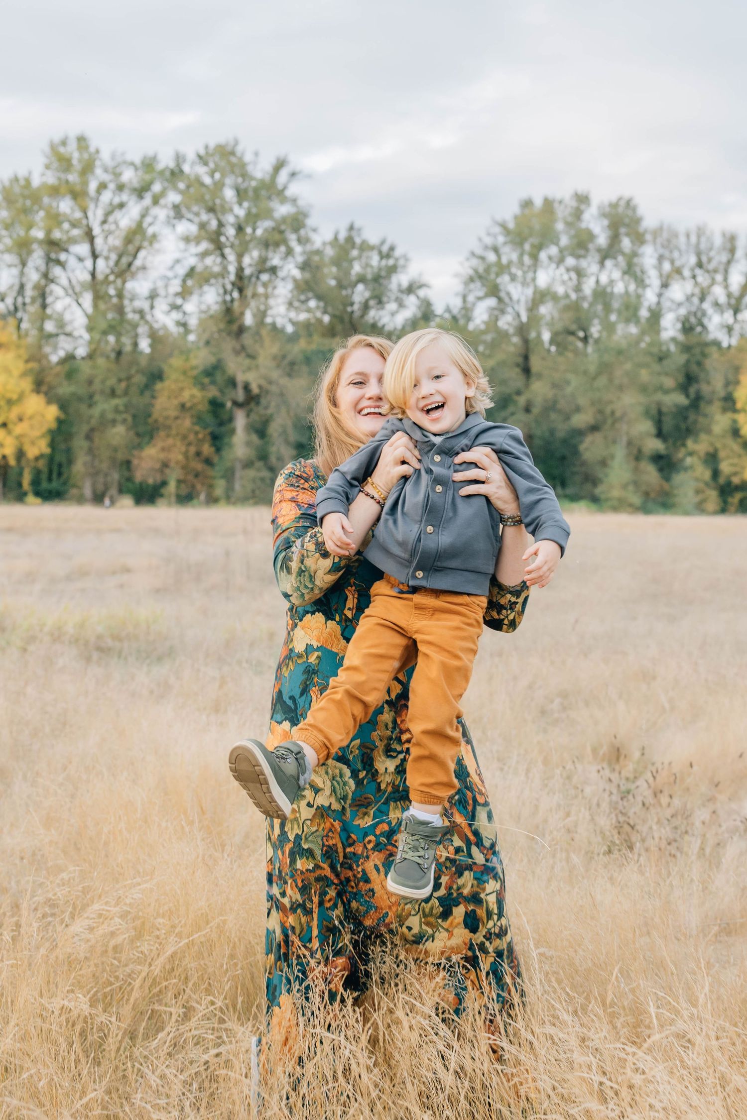 Laughing mother in floral dress lifting her young son in a golden autumn field.