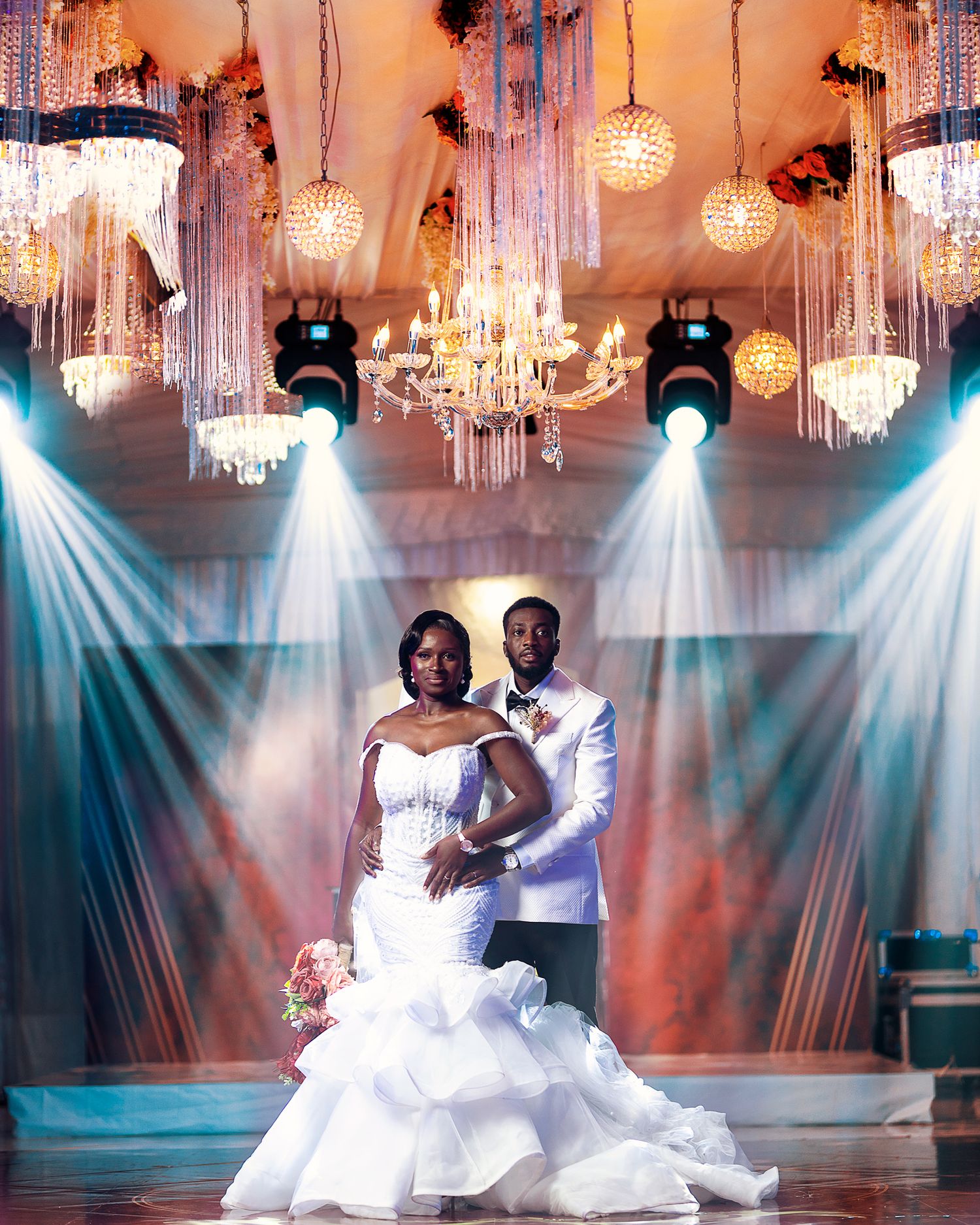 Dramatic wedding portrait under crystal chandeliers with spotlights creating atmospheric scene.
