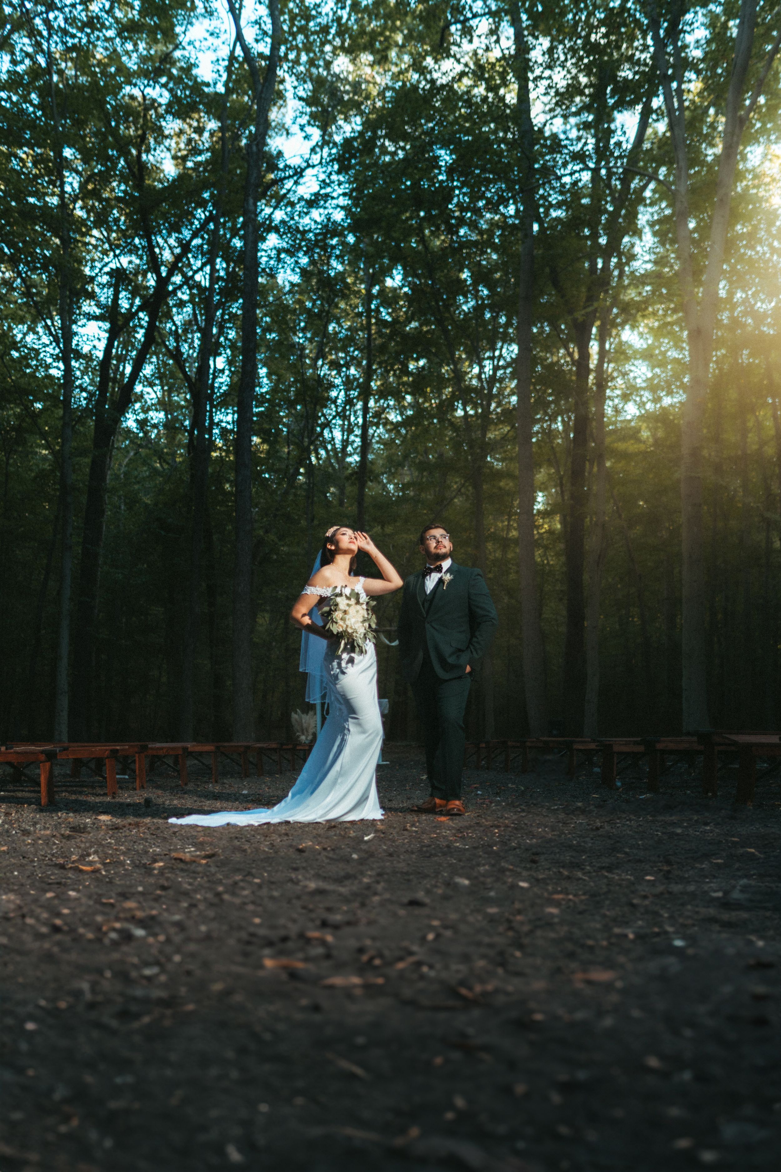 A couple in wedding attire poses in a forest clearing with sunlight streaming through tall trees at dusk.
