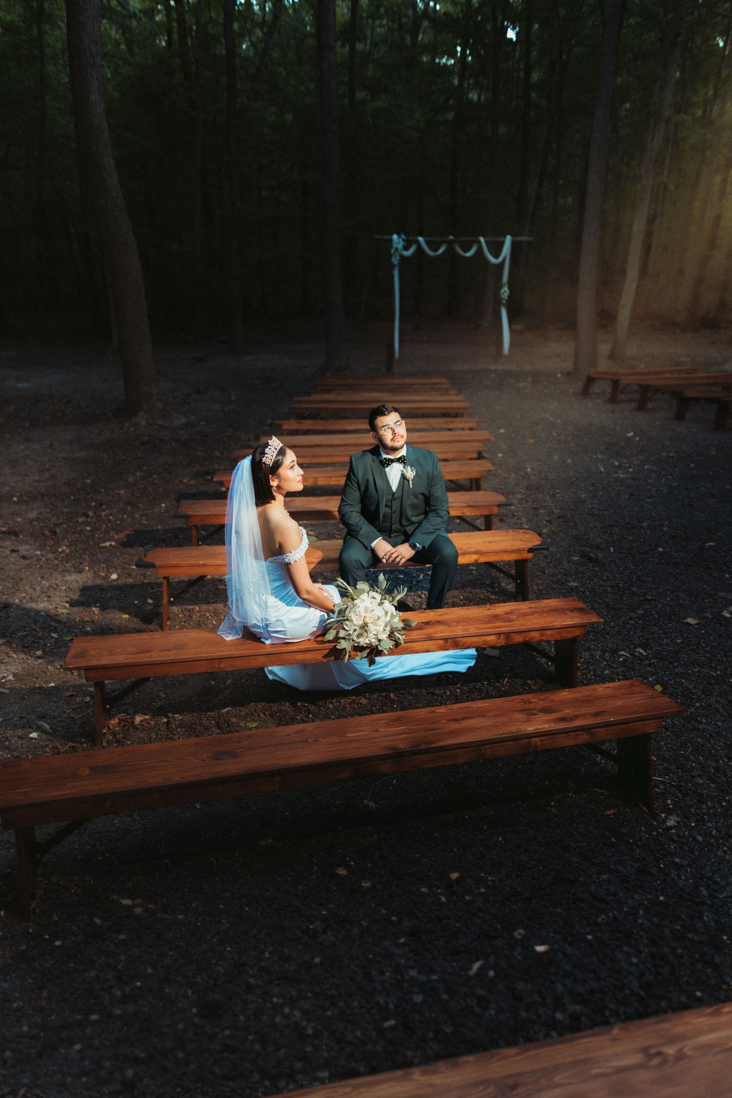 Rustic wooden bench seating in a forest creates an intimate wedding ceremony setting at dusk.