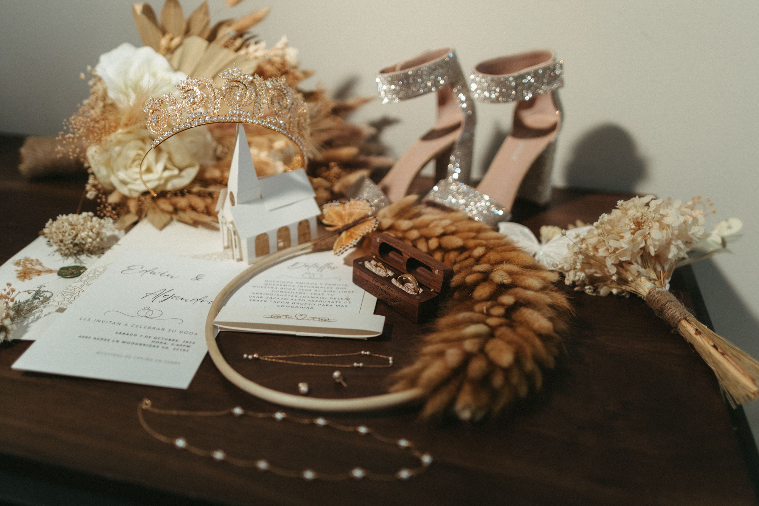 Wedding accessories including blush heels, invitations, and dried flowers arranged on dark wooden surface.