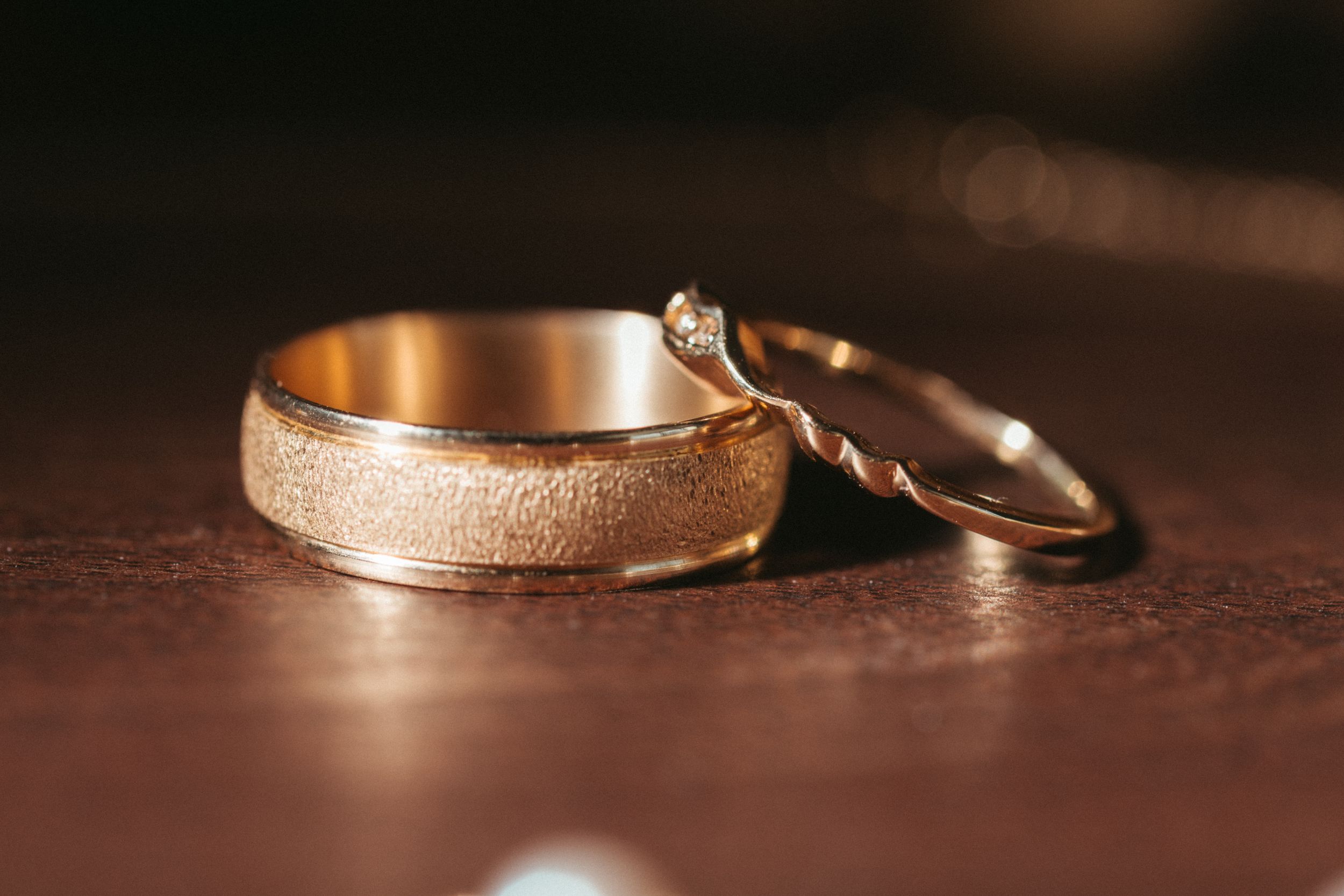 Gold wedding bands with textured and smooth finishes rest on dark wooden surface in warm lighting.