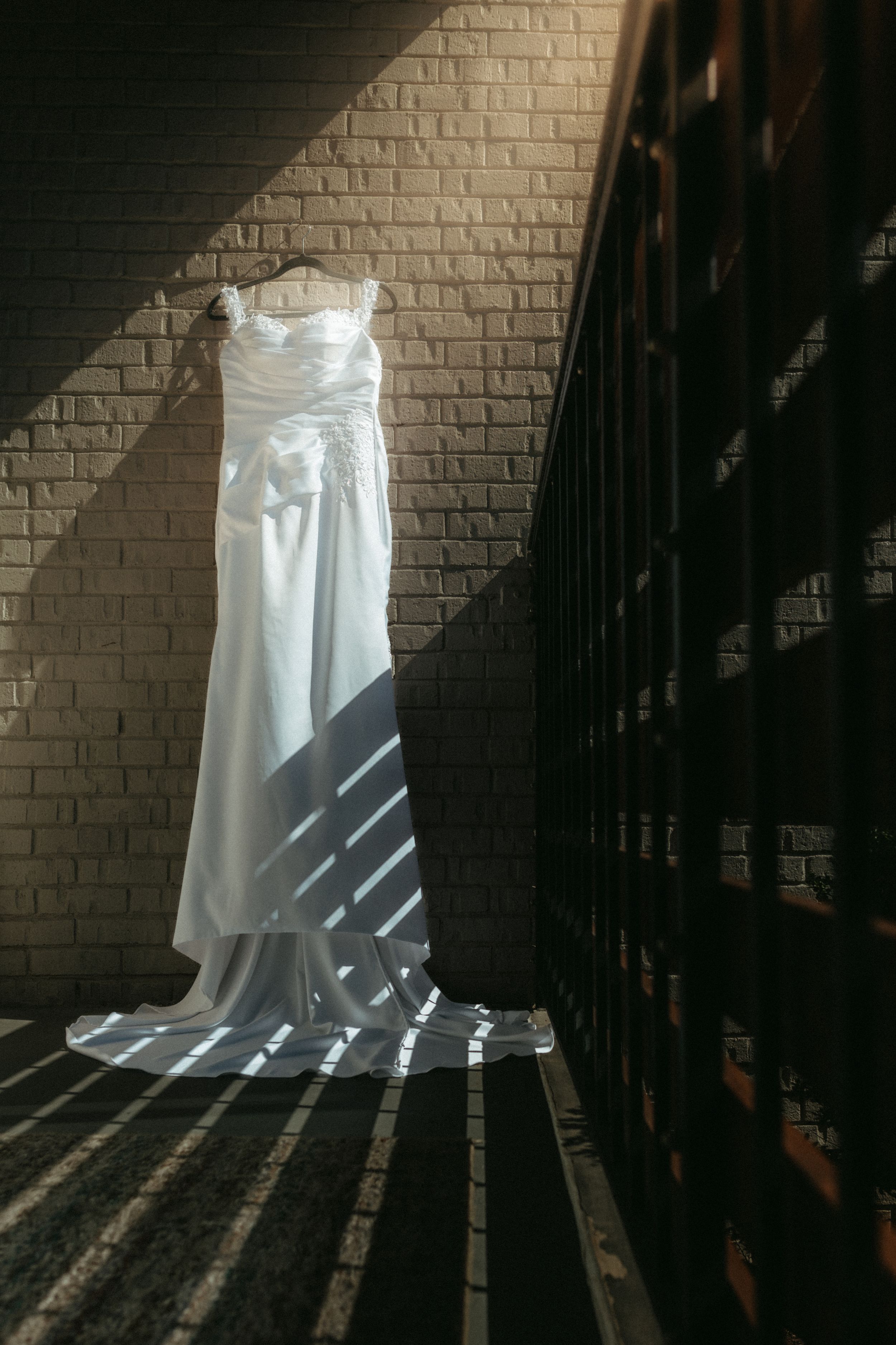 A white wedding dress hangs against a brick wall, casting dramatic shadows through a staircase railing.