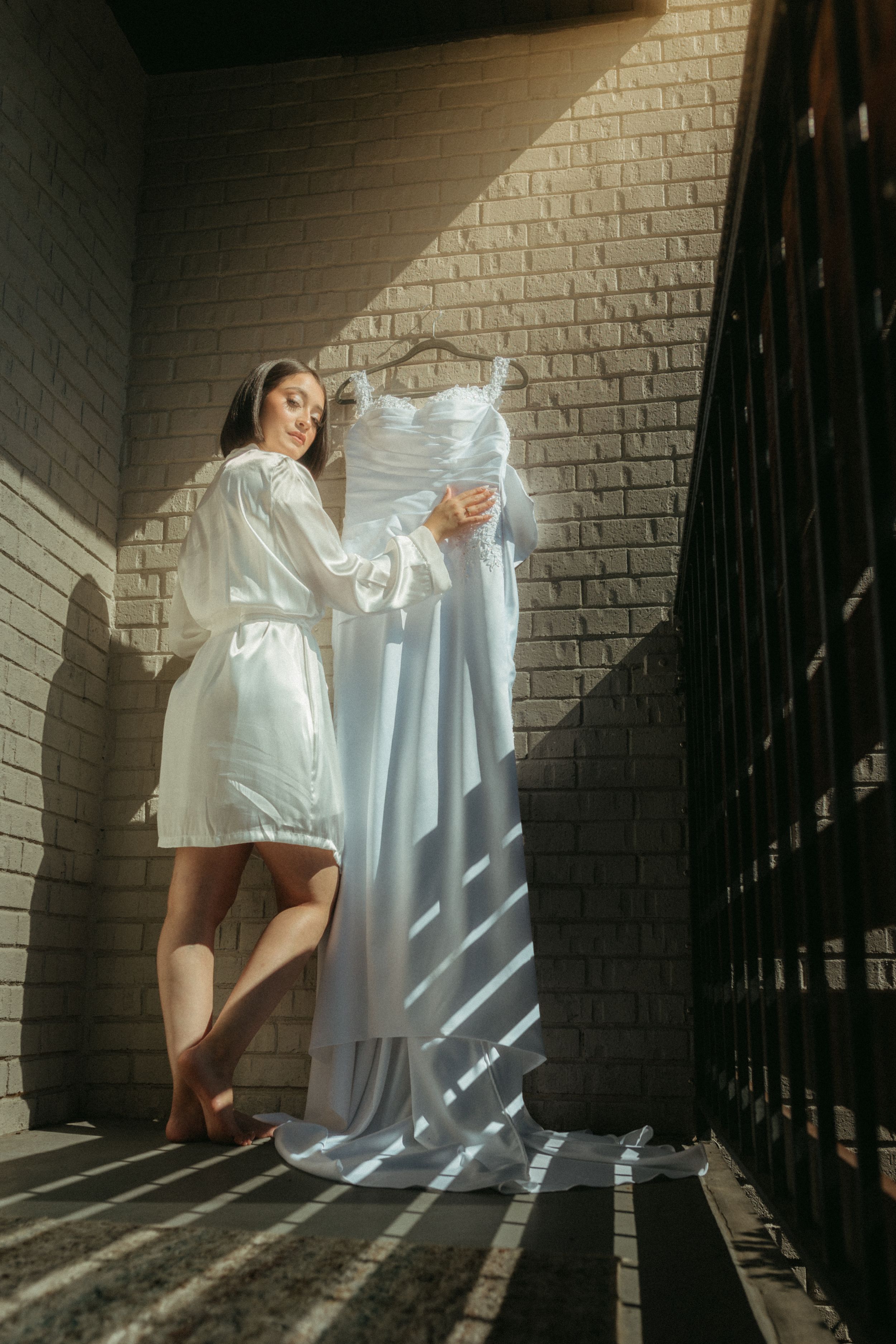 A person in a white silk robe stands holding a wedding dress against a brick wall with dramatic shadows cast through railings.