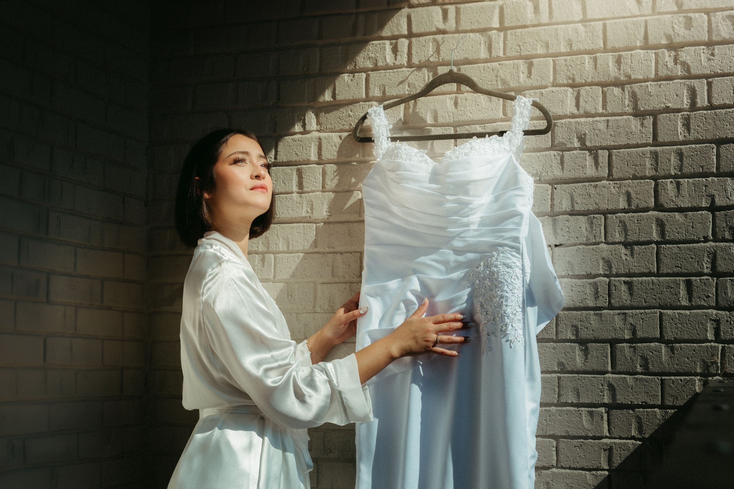 Person in white robe holds up elegant wedding dress on hanger against brick wall in preparation moment.