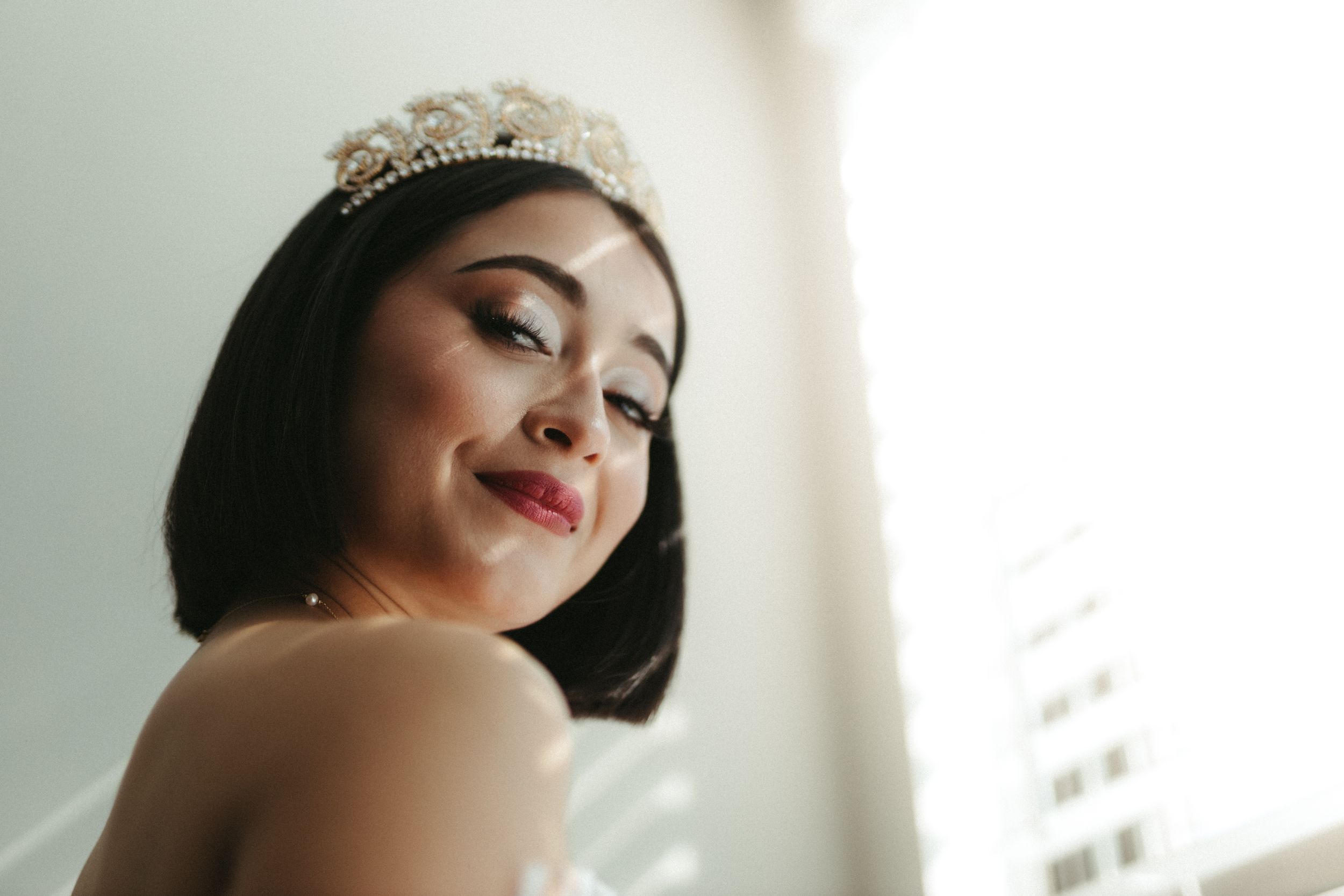 Elegant bride in a tiara smiles softly in a romantic natural light setting.