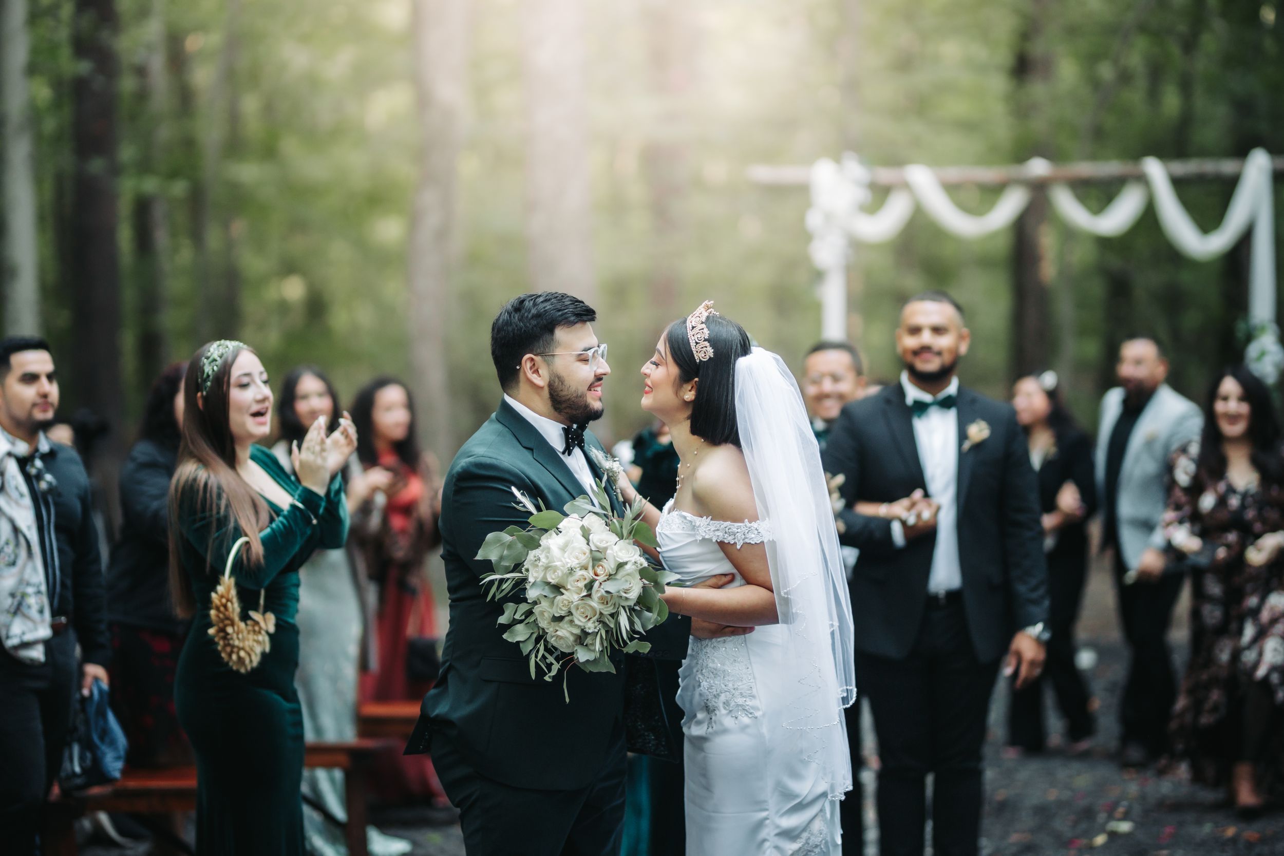 A couple celebrates their wedding ceremony in a lush forest setting surrounded by guests under hanging decorations.