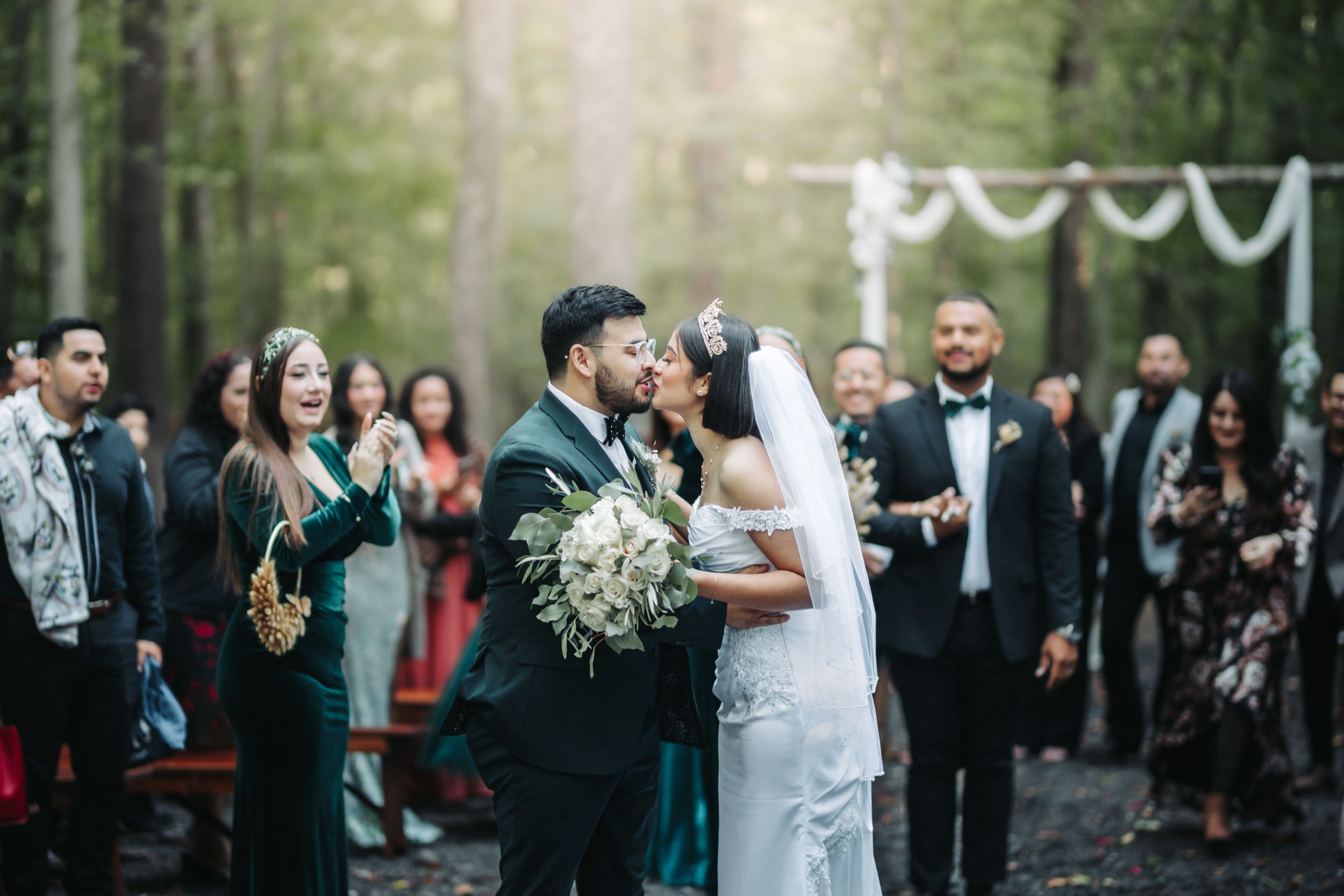 A romantic outdoor forest wedding ceremony with guests watching the couple share a kiss under white draped fabric decor.
