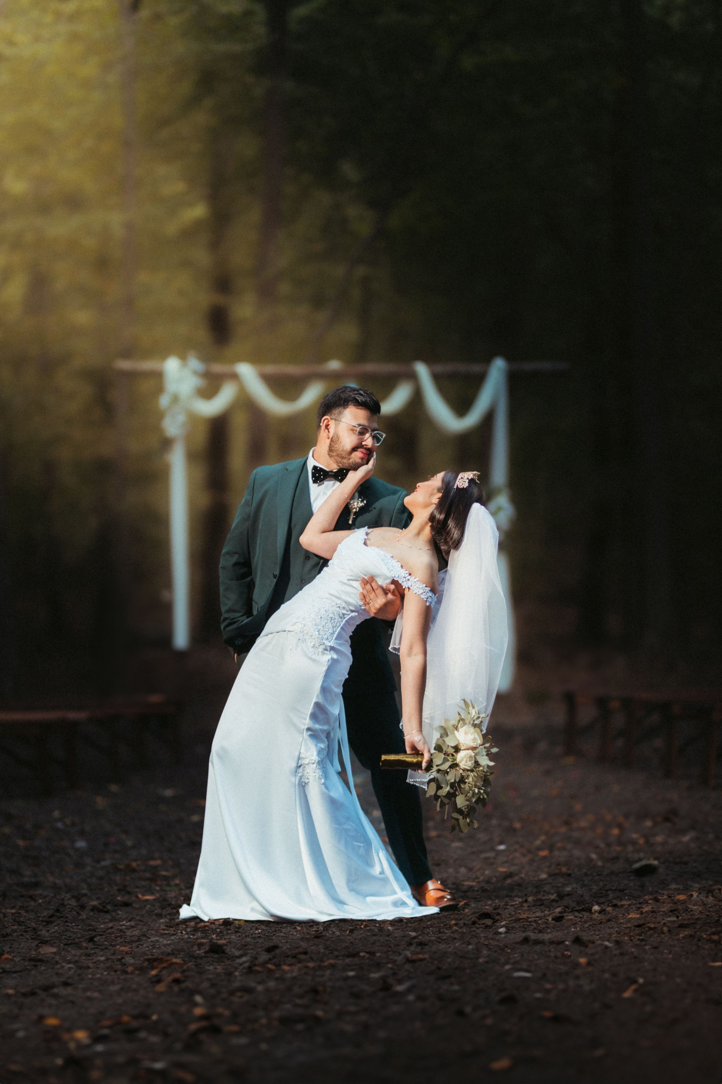 Couple in wedding attire pose romantically in a forest setting with twinkle lights in the background.