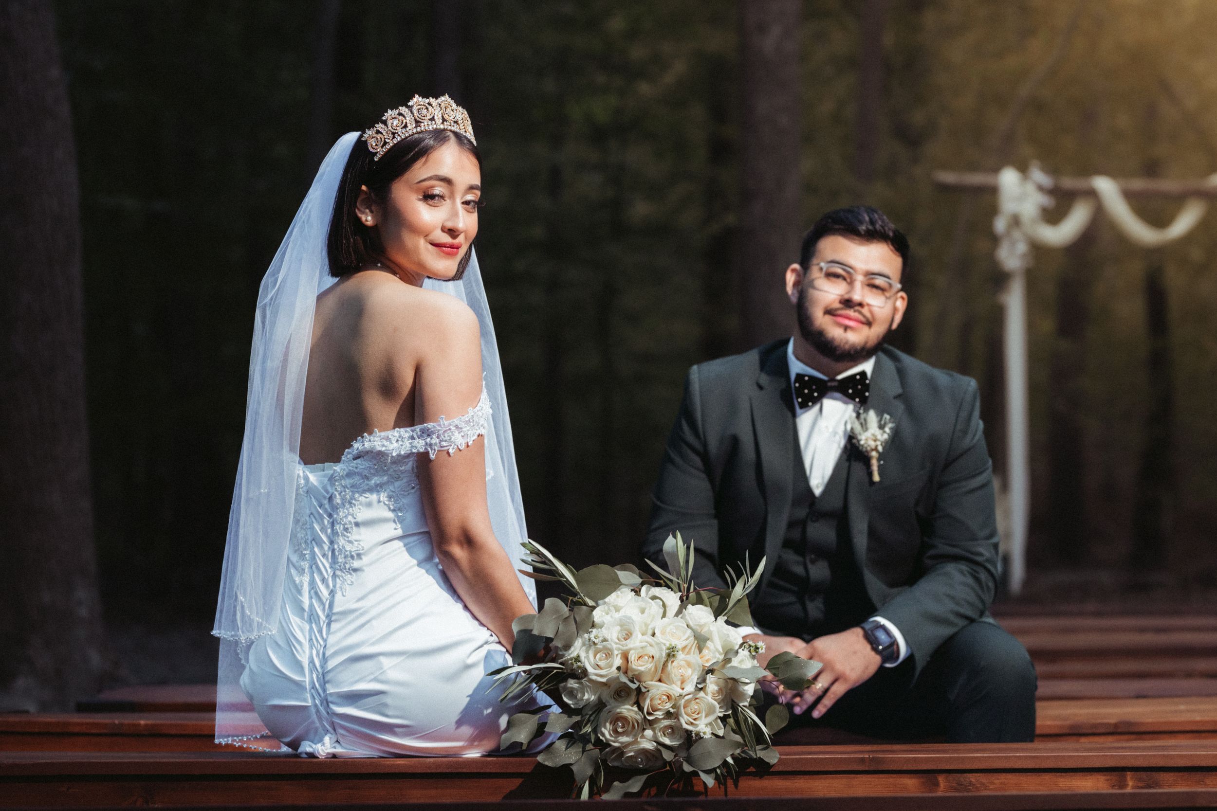 A bride in a white dress sits and turns on a wooden bench beside a groom in a black tuxedo during an outdoor wedding.