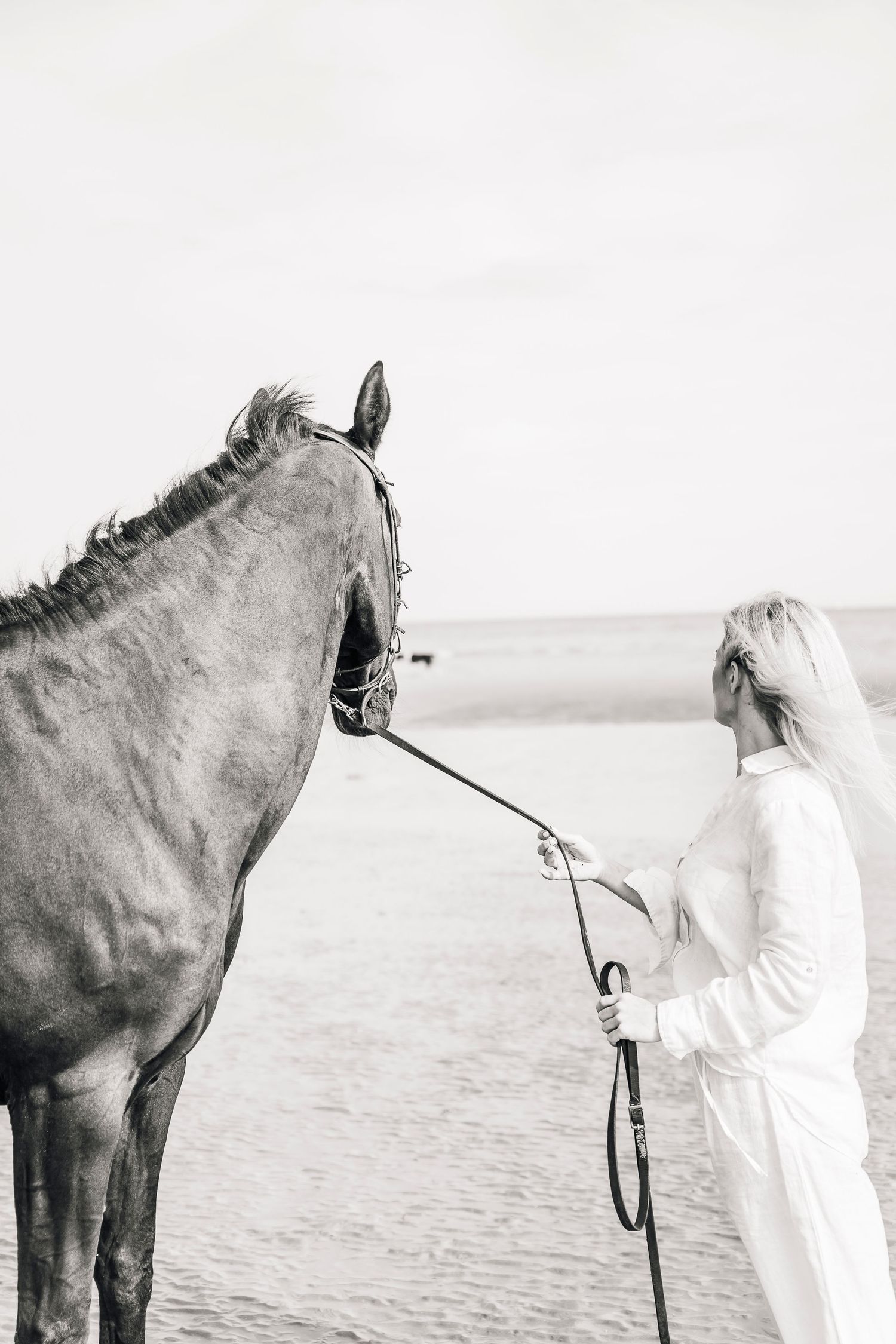 A person in white stands with a dark horse on a serene beach in black and white.