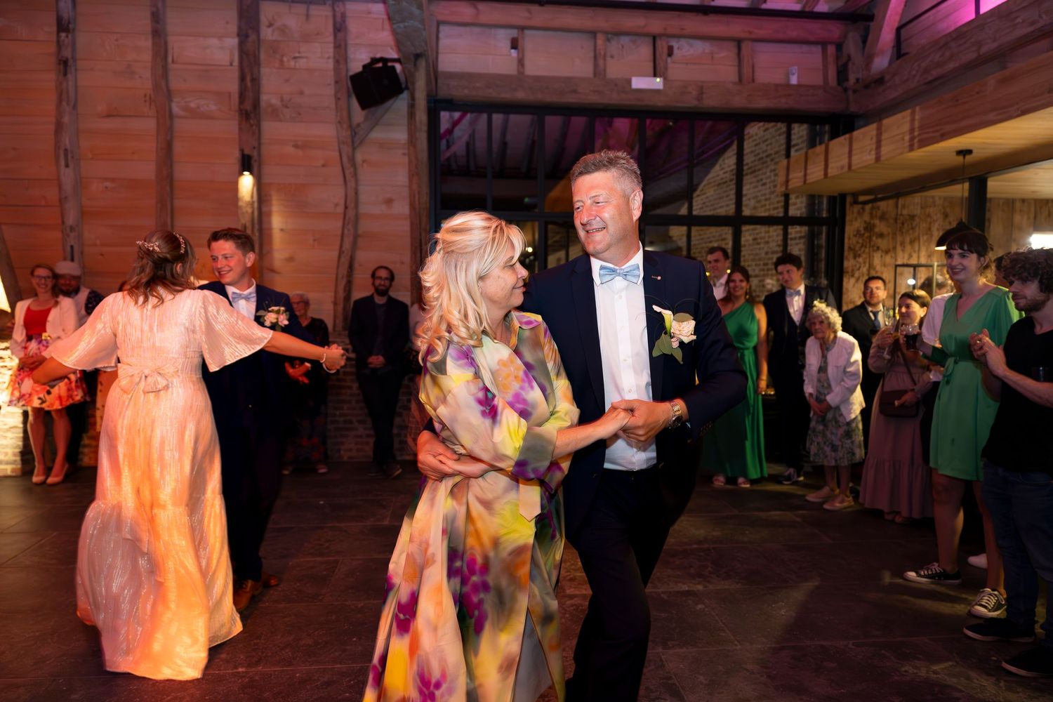 Guests dancing at a wedding reception in a rustic venue with exposed brick walls and colored lighting.