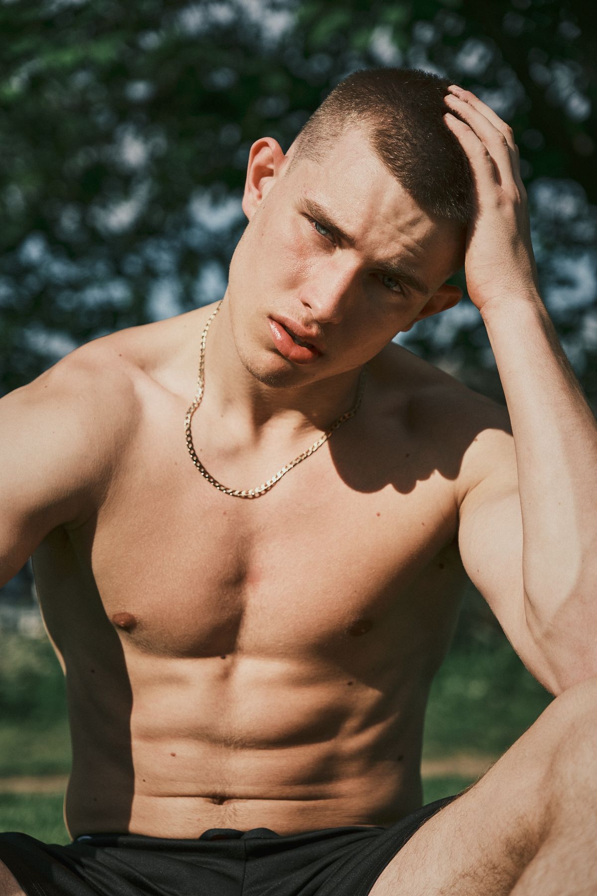 hirtless male model with short hair and defined abs, sitting outdoors in sunlight with focused expression