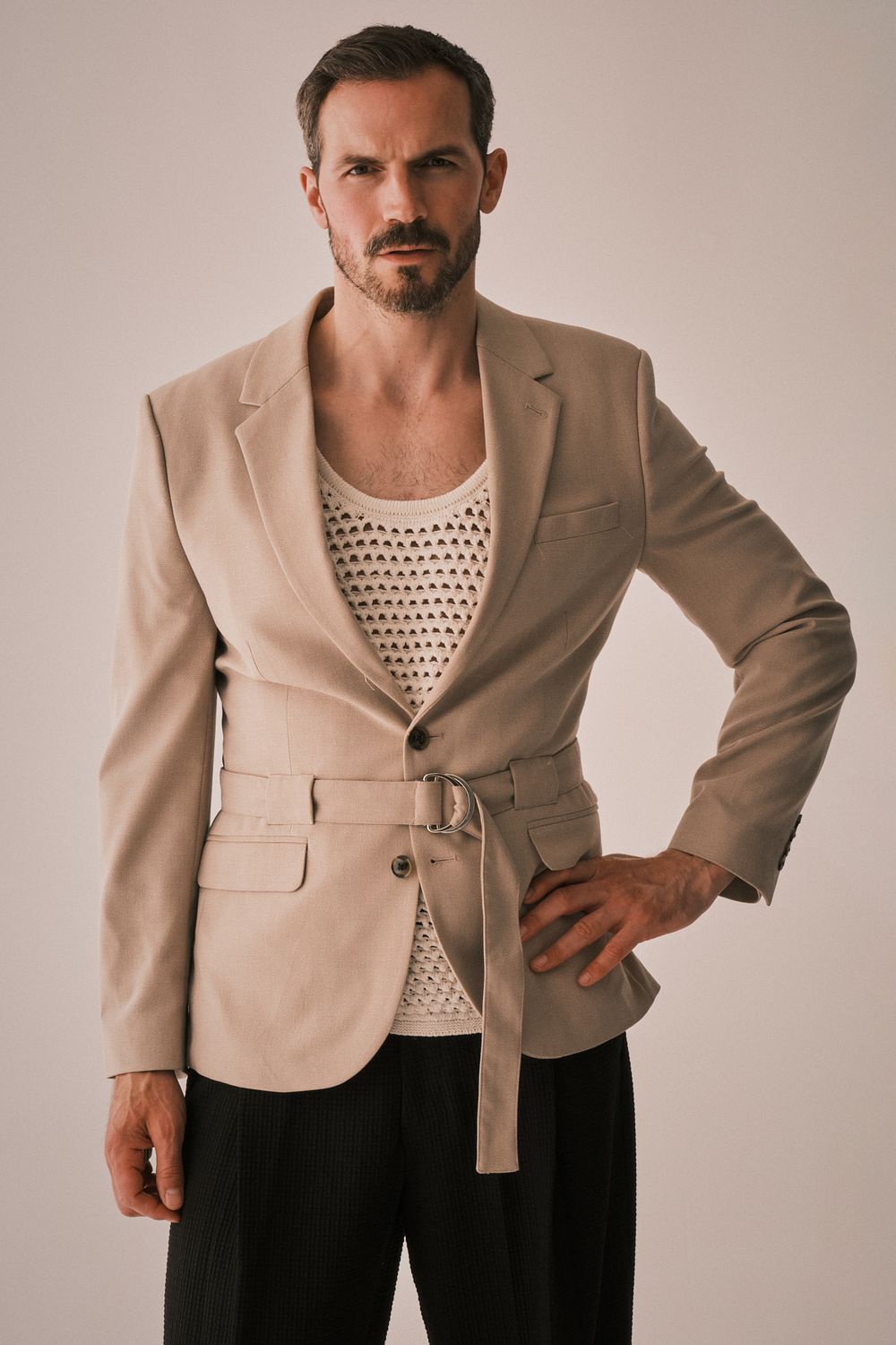 Male model in a belted beige blazer over a mesh vest, exuding a retro vibe against a clean, minimalist background
