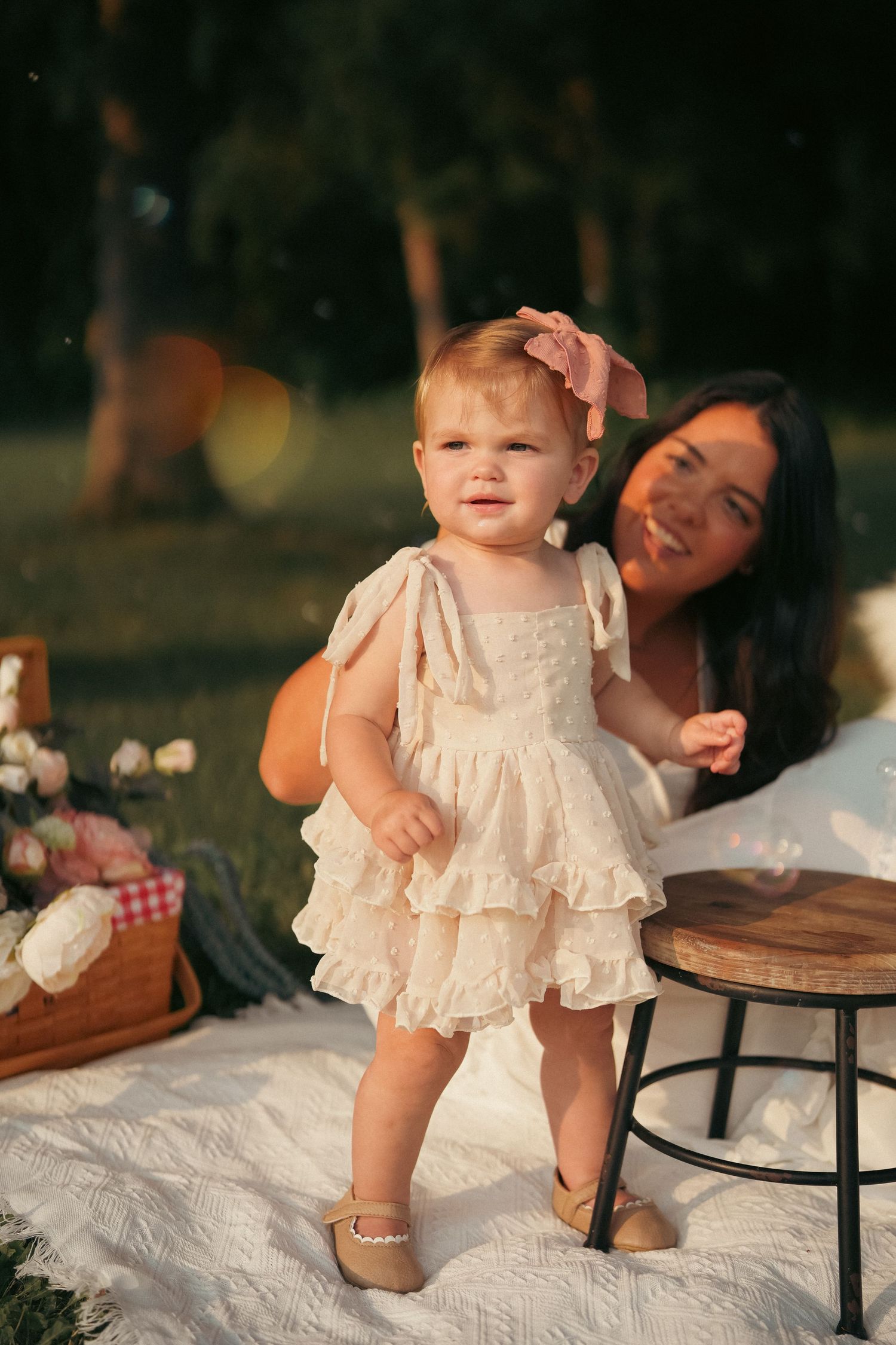 Baby in white lace dress sitting on picnic blanket outdoors.