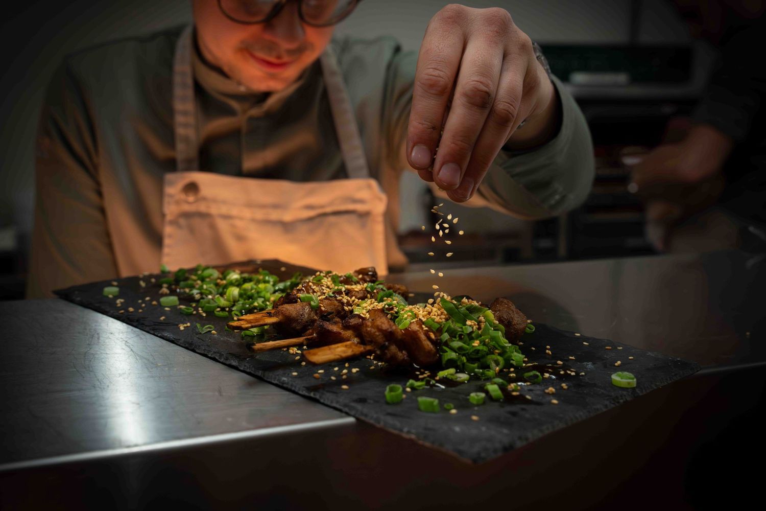 Chef finishing a meat dish with fresh herbs and seasonings in dramatic lighting.