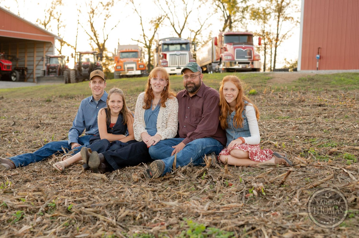 Family sitting together on the ground near a red barn and farm equipment during golden hour.